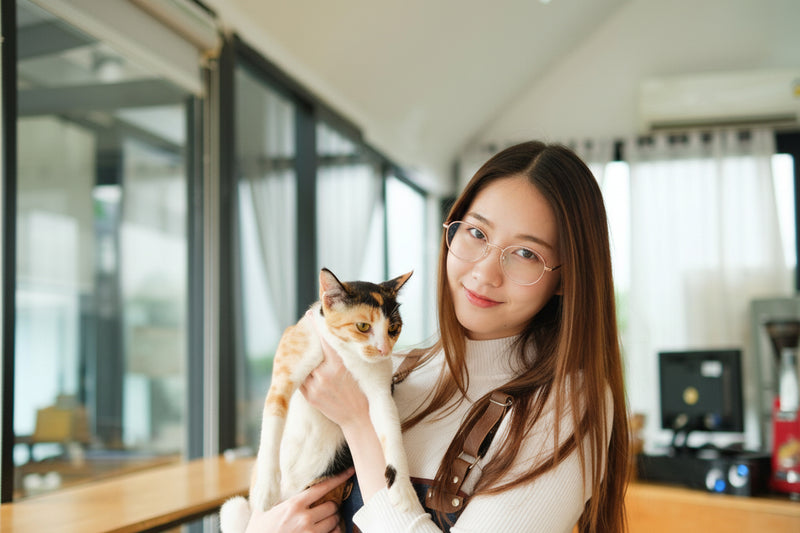 A woman with long brown hair and glasses holds a calico cat in an indoor setting with large windows.